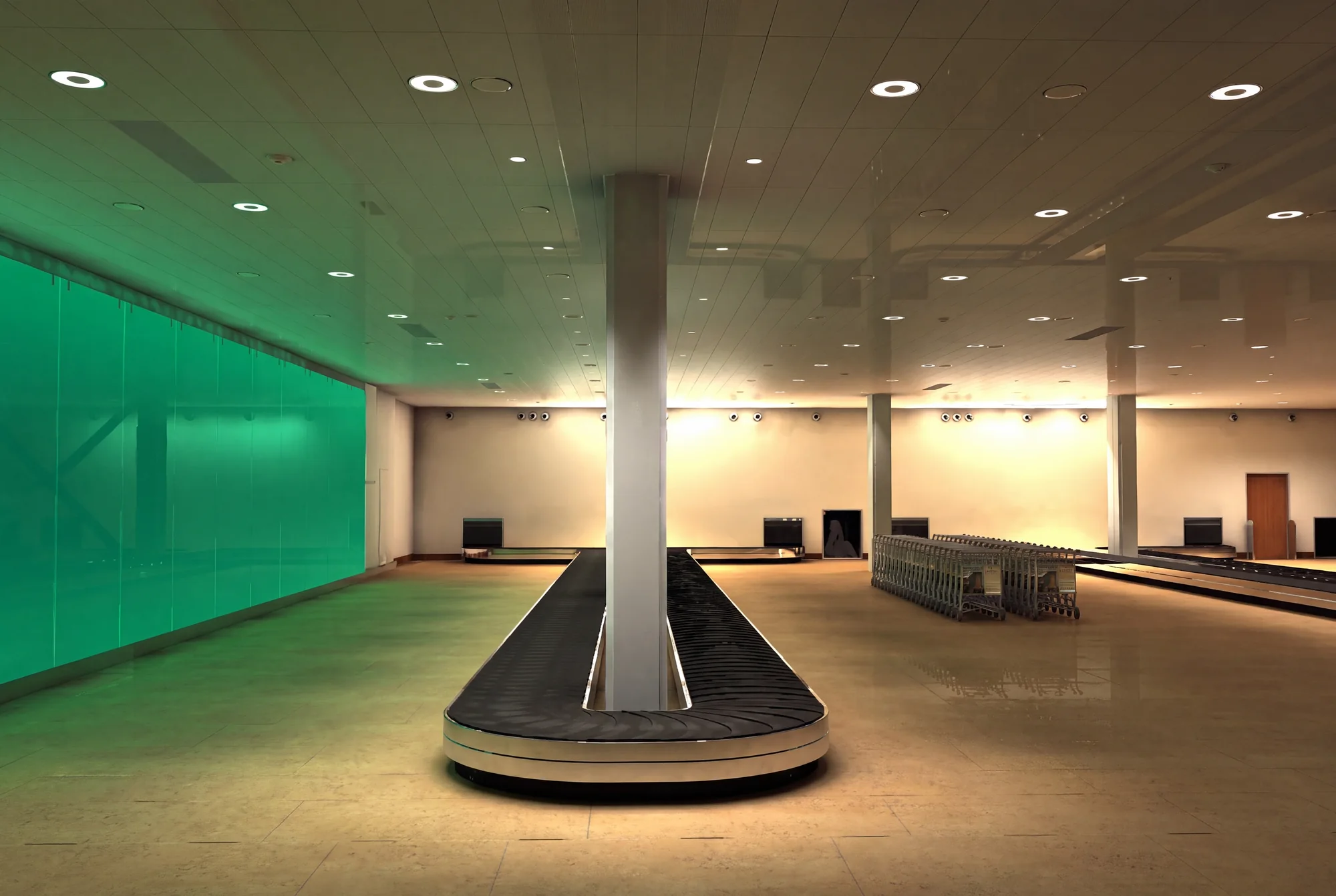 Empty airport arrivals hall, frontal view down the carousel with a glowing green glass wall on the left and stacked luggage trolleys