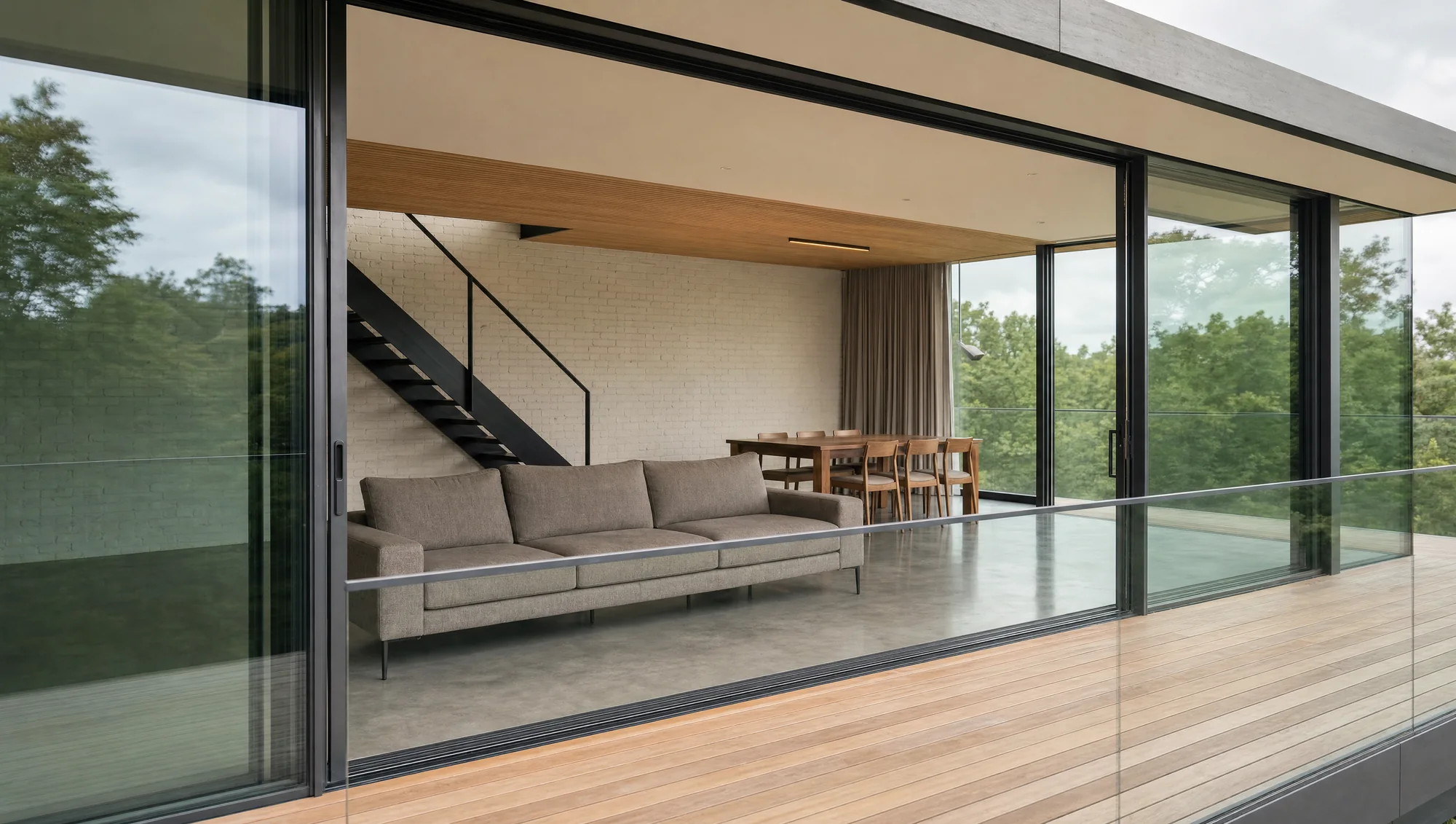 View from the terrace through the open sliding doors — sofa, black steel stair against a white brick wall, dining table beyond