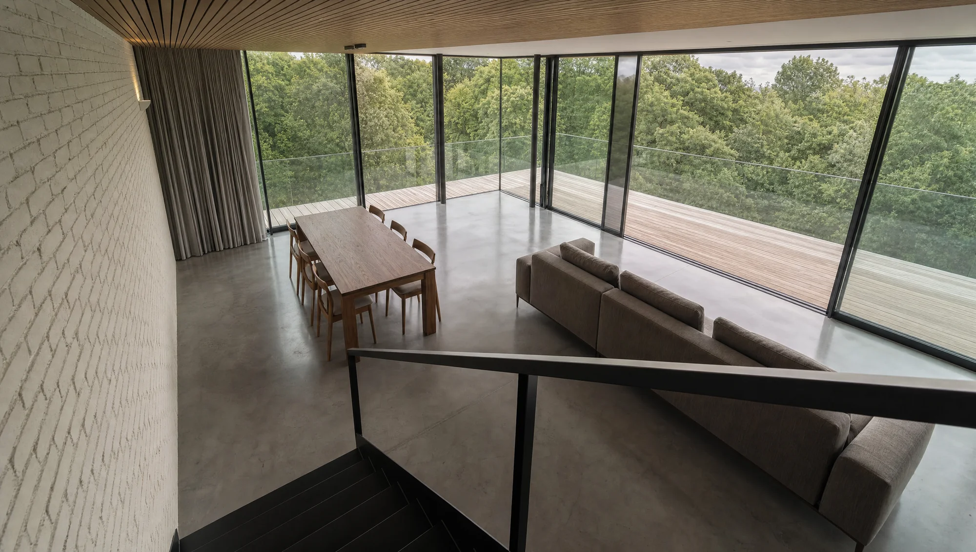 Overview from the stair landing — dining table, sofa and the full-height glazing onto the timber deck and treetops