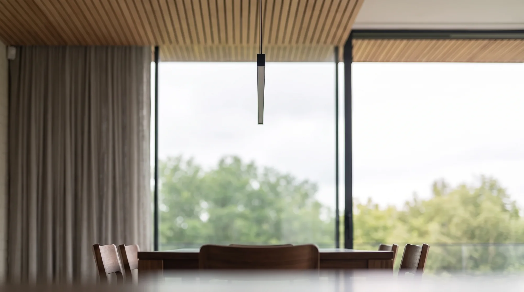 Slim pendant light over the table, linen curtain and treetops through the glass