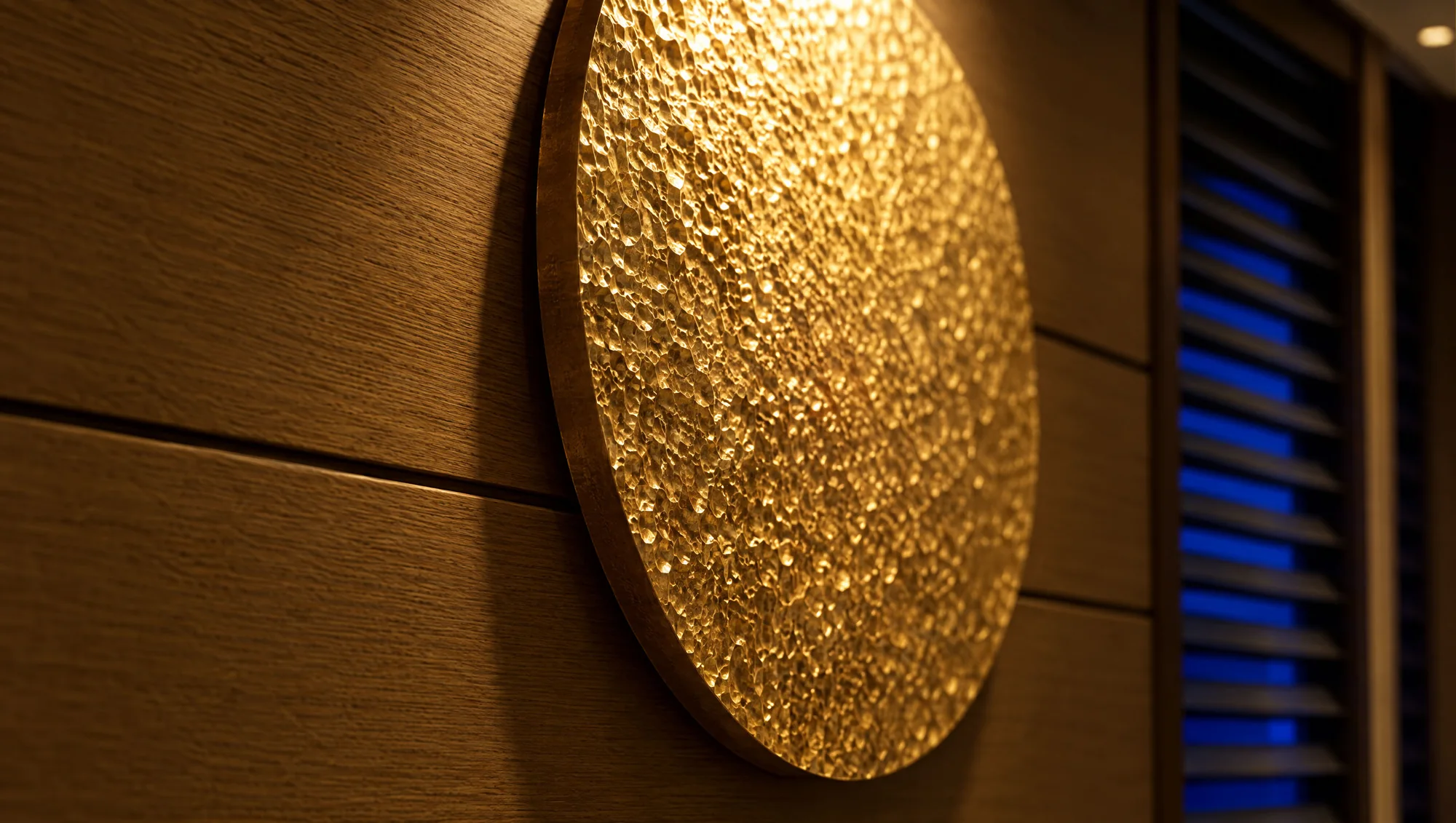 Close-up of the hammered gold wall disc on oak panelling, blue night light through the louvres