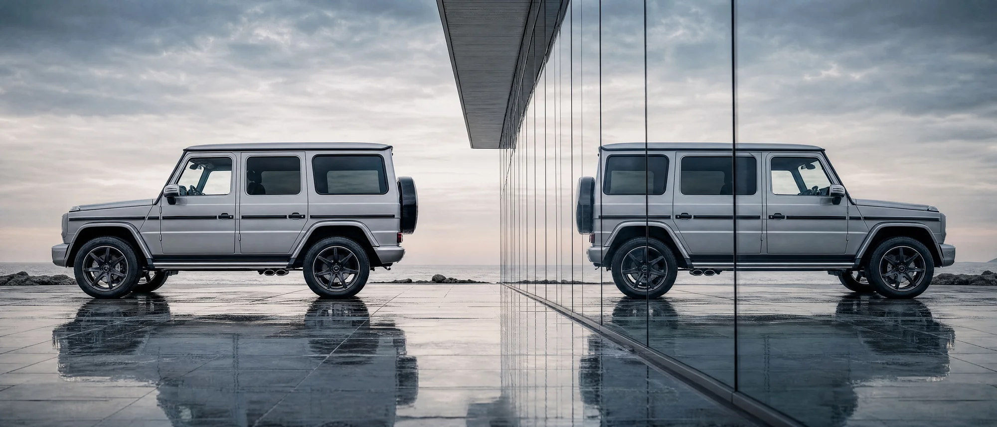 Silver G-Class beside a mirrored glass building facade, coastal overcast sky