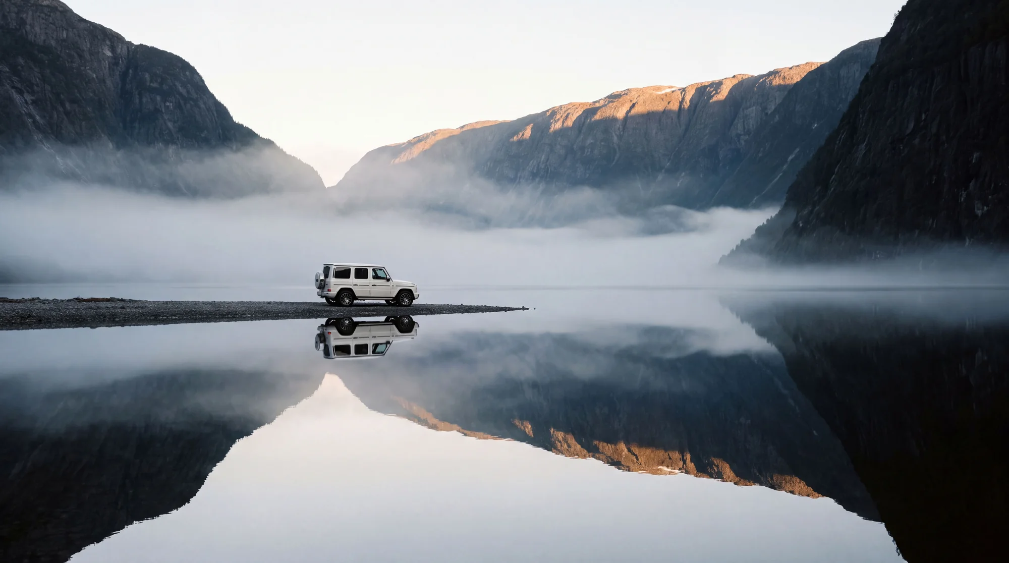 White G-Class on a causeway in a foggy fjord landscape, mountains reflected in still water