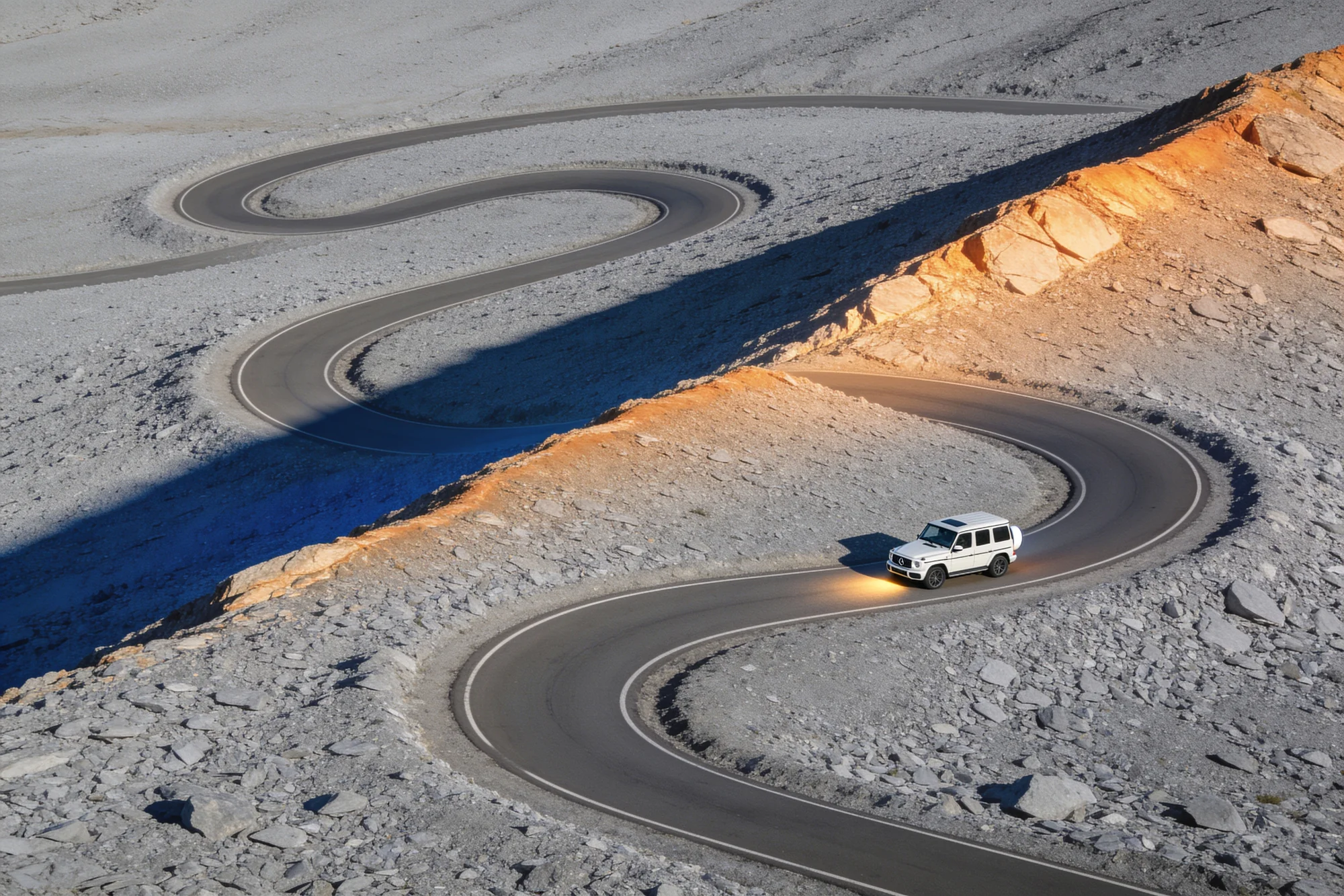 Aerial view of a white G-Class with headlights on, driving a winding mountain road