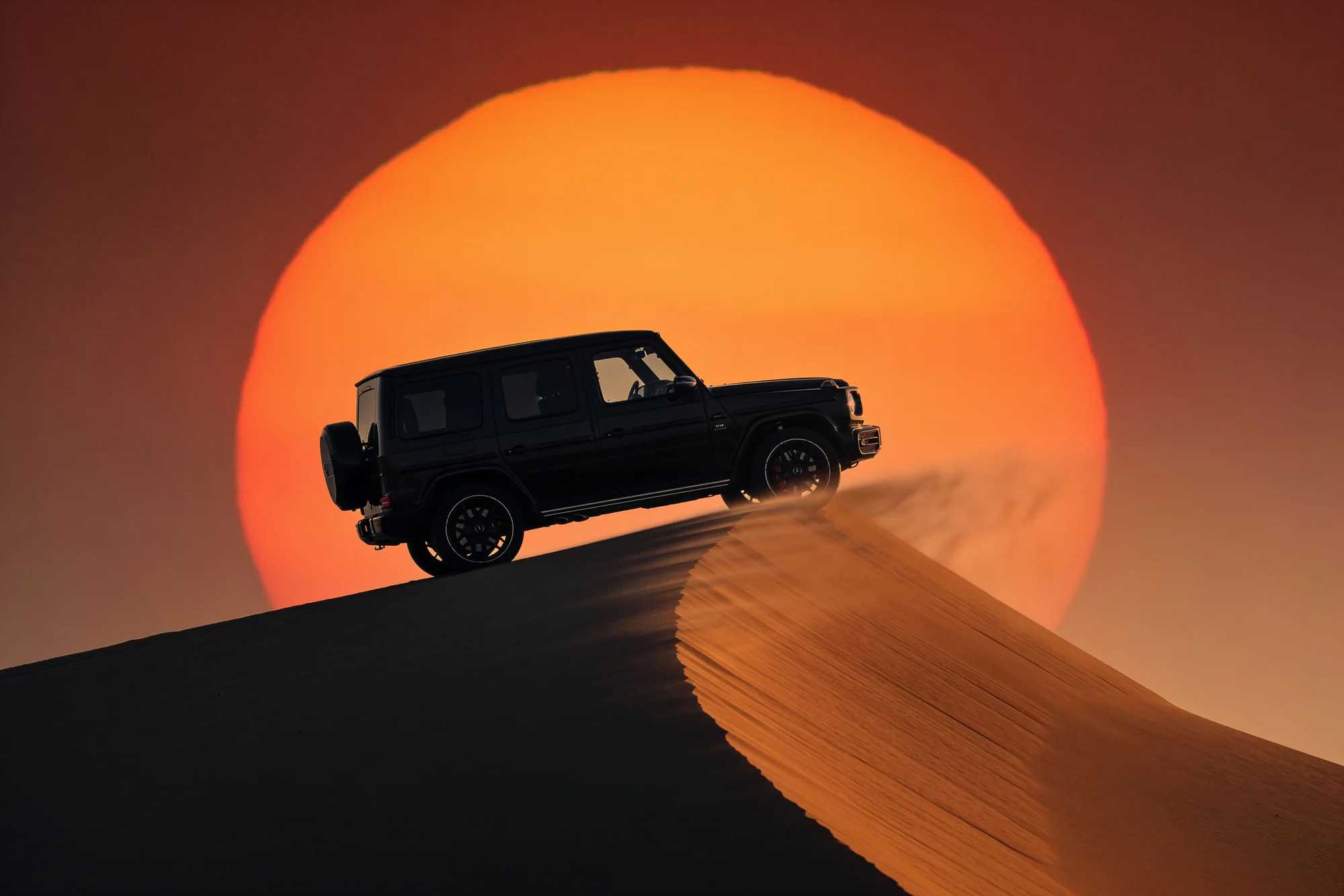 Black G-Class silhouette cresting a sand dune against a huge setting sun